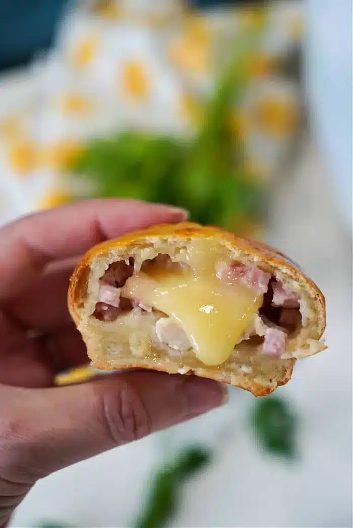 A hand holds a baguette piece filled with melted cheese and small diced ham, reminiscent of the flavors in an air fryer chicken cordon bleu. A green herb is blurred in the background.