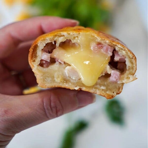 A hand holds a baked bread roll reminiscent of air fryer chicken cordon bleu, filled with melted cheese and small pieces of ham. A bite reveals the gooey interior, while greenery and other ingredients blur in the background.