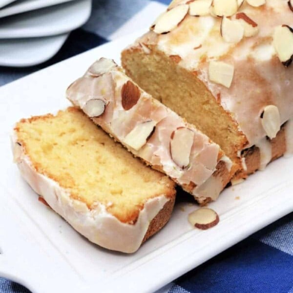 Copycat Starbucks lemon loaf cake with icing and almond slices on a white platter, placed on a blue checkered tablecloth.