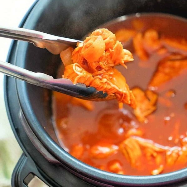 Tongs grasping tender instant pot shredded chicken in a pressure cooker brimming with rich red sauce.