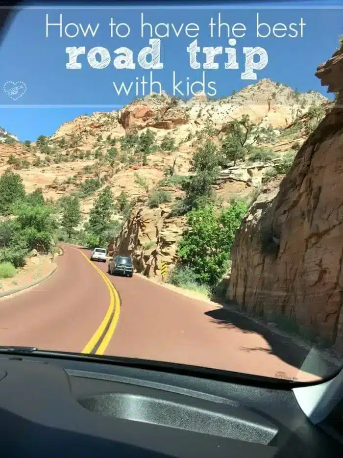 A scenic view from a car dashboard window reveals a winding road through rocky, mountainous terrain under a clear blue sky. Two cars appear ahead, perfect for a game of "I Spy." Text overlay reads, "How to have the best road trip with kids.