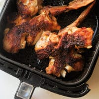 Close-up of grilled turkey wings inside an air fryer basket. The wings appear well-cooked with a seasoned, crispy exterior. The air fryer is black with a clear handle at the front.