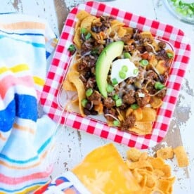 A plate of nachos topped with avocado, sour cream, and green onions sits next to a Fritos bag and colorful napkin, reminiscent of an Instant Pot Frito chili pie recipe.