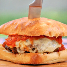 A New York pizza burger with melted cheese and tomato sauce on a bun, topped with a knife, sits enticingly on a wooden board.