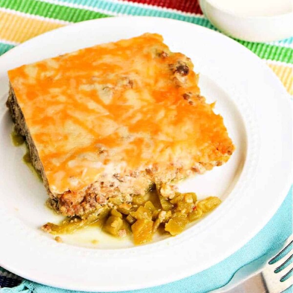 A slice of cheesy chile relleno casserole with ground meat and green chilies sits invitingly on a white plate, complemented by a colorful placemat underneath.