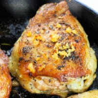 Close-up of a seasoned, golden-brown garlic butter chicken thigh in a skillet, topped with minced garlic and herbs.