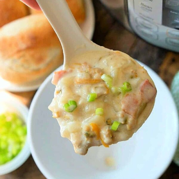 A spoon is held over a bowl, filled with an easy Instant Pot clam chowder, boasting chunks of meat and vegetables, garnished with chopped green onions. Bread rolls are blurred in the background.