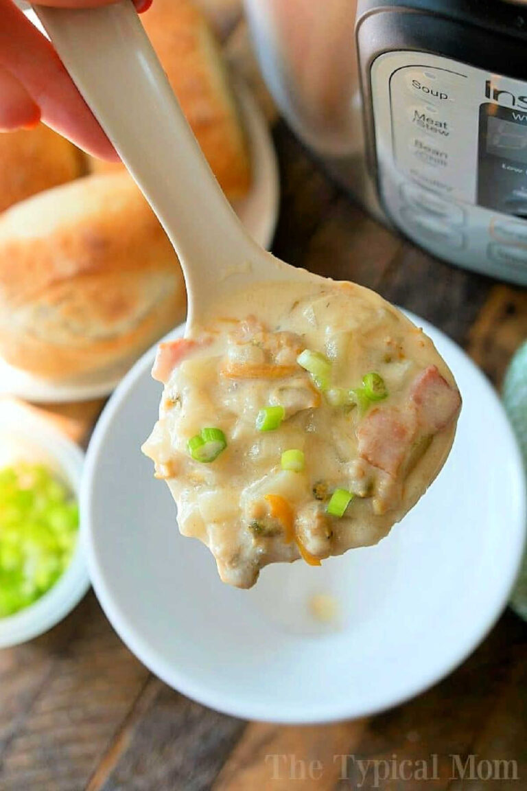 A spoon is held over a bowl, filled with an easy Instant Pot clam chowder, boasting chunks of meat and vegetables, garnished with chopped green onions. Bread rolls are blurred in the background.