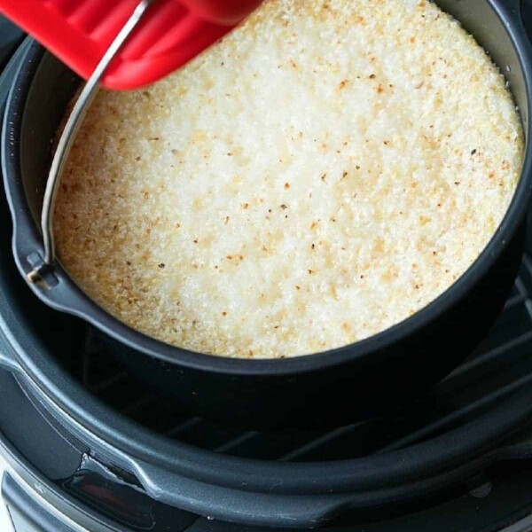 A red oven mitt lifts a pan of golden-browned, baked dish from a black pressure cooker, as if serving up warm Instant Pot grits.