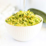 A white bowl brimming with spiralized zucchini noodles, perfectly crisped in the air fryer, sits invitingly with a cucumber in the background.