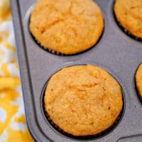 A close-up of a muffin tray reveals six golden-brown pumpkin banana muffins nestled in paper liners. Made with cake mix, these treats boast a perfect crust. A yellow and white patterned cloth peeks from beneath the tray, adding a touch of color.