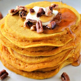 A stack of pumpkin pancakes, one of the easy pumpkin desserts, topped with butter, pecans, and drizzled maple syrup sits on a plate. A few pecans are scattered around the plate. The pancakes have a light golden-brown color.