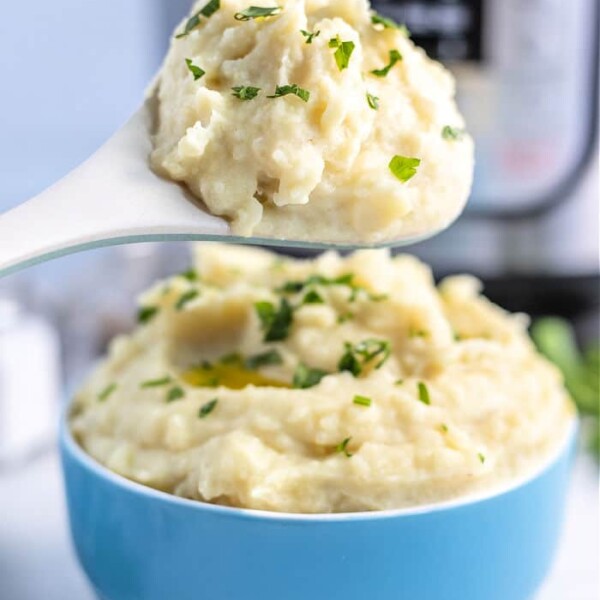 A spoonful of garlic mashed potatoes garnished with chopped parsley is held above a blue bowl filled with more mashed potatoes. A pressure cooker is visible in the background.