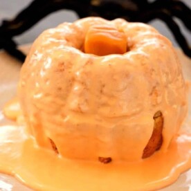 A delightful mini bundt cake made with pumpkin cake mix, topped with orange icing and a caramel, all elegantly presented on a plate with a decorative spider behind.