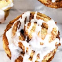 Close-up of a cinnamon roll muffin topped with white icing. The golden brown treat reveals its swirled layers, dusted with sprinkled cinnamon, as the icing drips down the sides. Two partial muffins are visible in the background on a marble surface.