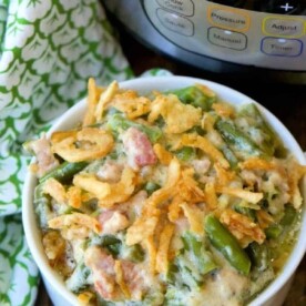 A bowl of instant pot green bean casserole topped with crispy onions sits beautifully in front of a pressure cooker, with a green patterned cloth nearby.