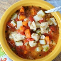 A yellow bowl of Instant Pot chicken vegetable soup brimming with diced potatoes, carrots, tomatoes, and peas, with a spoon resting inside. The bowl is placed on a decorative cloth.