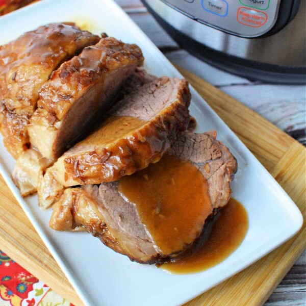 Sliced roast beef with gravy on a white plate next to an Instant Pot on a wooden cutting board. The Instant Pot rump roast appears tender and juicy.