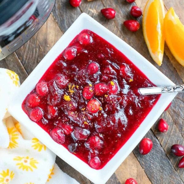 A square bowl of instant pot cranberry sauce sits on a wooden table, adorned with orange slices, cinnamon sticks, and fresh cranberries.