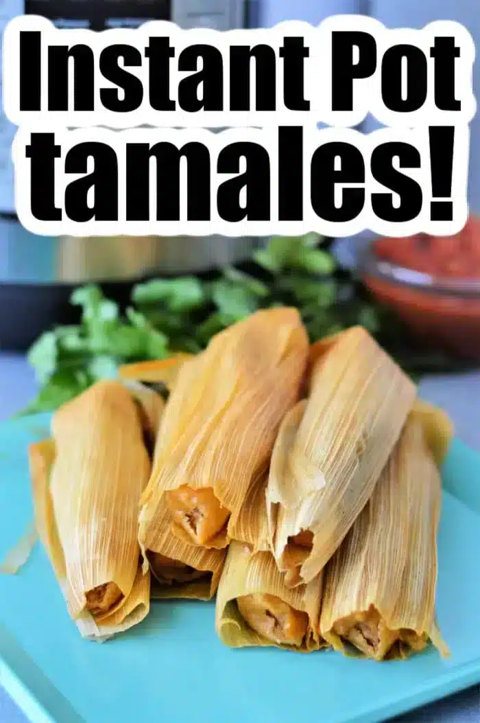 A stack of tamales wrapped in corn husks on a blue surface. The text "Steam tamales in Instant Pot!" is prominently displayed above. In the background, there is a blurred image of cilantro and a bowl.