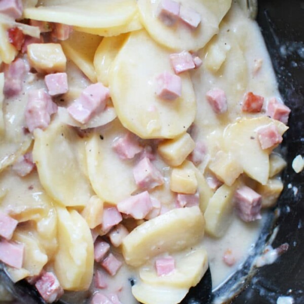 Close-up of crockpot ham and potatoes featuring sliced potatoes, diced ham, and onions in a creamy sauce. A black spoon rests in the mixture, perfect for stirring or serving this hearty dish.