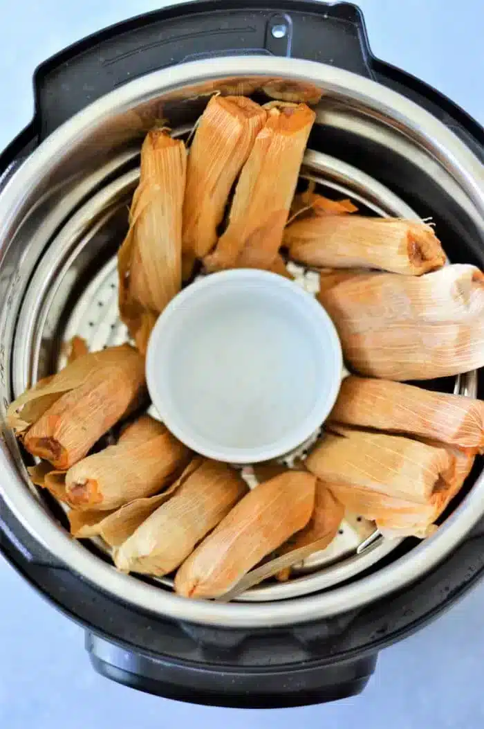 A circular arrangement of tamales with corn husks is expertly placed to steam in the Instant Pot, encircling an empty white cup in the center. The metal interior of the pot gleams, ready for a savory transformation.