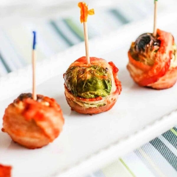 Three crispy Brussels sprouts with bacon, held together by toothpicks, stand in a row on a white rectangular plate, arranged on a striped surface.