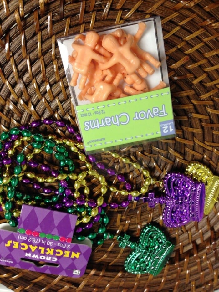 A wicker basket holds colorful beaded necklaces in purple, green, and gold with crown and heart-shaped pendants. Nestled among them is a box of small plastic baby figurines labeled Favor Charms—perfect for topping a Mardi Gras cake.