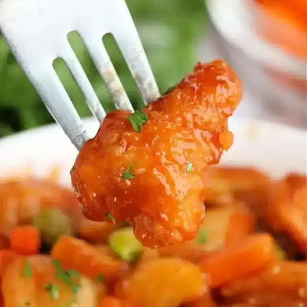 A fork holds a piece of orange-glazed chicken, perfectly crispy from the air fryer, over a bowl filled with more pieces and mixed vegetables like carrots and peas. Parsley is sprinkled on top for garnish.