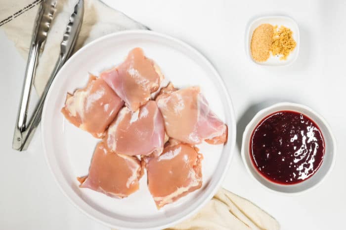 Raw chicken thighs rest on a plate, ready to transform into slow cooker BBQ perfection. Nearby, tangy sauce and savory seasonings await their turn, next to a pair of tongs resting on a cloth.