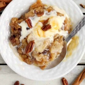 A creamy crockpot apple dump cake dessert, topped with pecans and served with a spoon on a white plate, surrounded by cinnamon sticks.