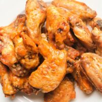 A plate of crispy, baked chicken wings is coated in a glossy orange sauce, arranged in a tempting pile. The lightly charred wings are served on a white dish, demonstrating an expert touch on how to bake frozen chicken wings to perfection.