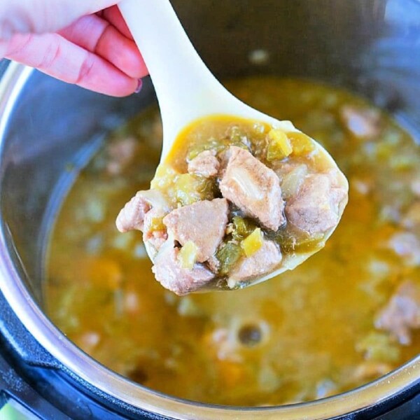 A ladle filled with a hearty stew containing chunks of meat and vegetables is being held above a pot. The stew, reminiscent of pressure cooker chili verde with its thick, brothy consistency, fills the pot halfway. A hand with painted nails holds the ladle.