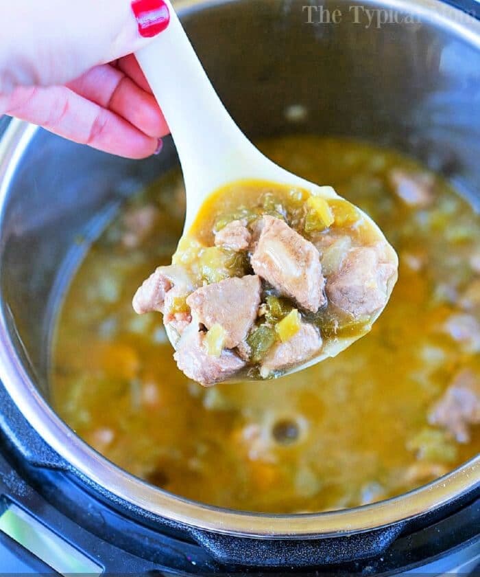 A ladle filled with a hearty stew containing chunks of meat and vegetables is being held above a pot. The stew, reminiscent of pressure cooker chili verde with its thick, brothy consistency, fills the pot halfway. A hand with painted nails holds the ladle.
