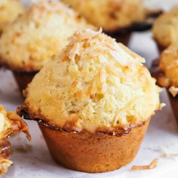 A close-up of a golden-brown coconut muffin crowned with toasted coconut flakes on a parchment-lined surface.