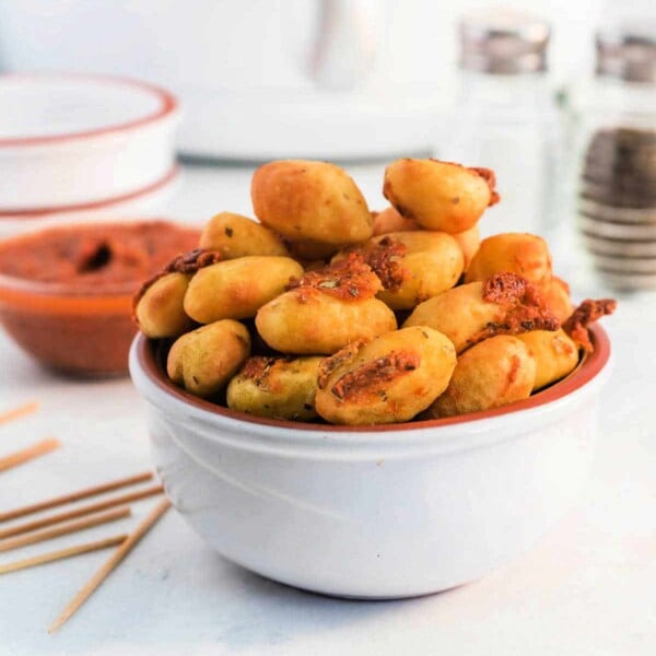 A white bowl filled with golden, air fryer gnocchi sits on the table, offering a crispy delight. Toothpicks are nearby for easy snacking, and condiments and seasoning shakers provide flavorful options in the background.