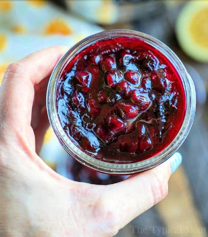 A hand demonstrating how to can jam holds a jar of homemade blackberry jam, showcasing the visible fruit pieces.