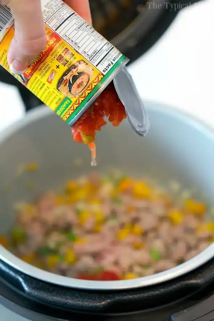 A hand pours rotel from a can into a slow cooker filled with ground meat, diced yellow bell peppers, and green vegetables for a hearty Mexican casserole with Rotel.