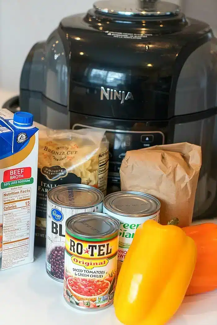 An assortment of kitchen items in front of a black Ninja pressure cooker: beef broth carton, bag of pasta, brown paper bag, canned black beans, diced tomatoes with green chilies, canned peas, and a yellow bell pepper—perfect for whipping up a spicy Mexican casserole.