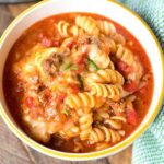 A bowl of rotini pasta, dubbed Ninja Foodi taco pasta, is topped with a chunky tomato and ground meat sauce, garnished with melted cheese and herbs. The bowl rests on a wooden surface beside a green cloth, with part of a kitchen appliance visible in the background.