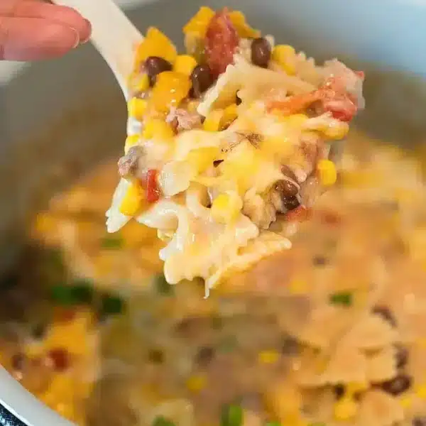 A spoonful of bowtie pasta mixed with corn, black beans, and melted cheese is held above the pressure cooker, showcasing a creamy Mexican casserole with diced tomatoes and green onions.