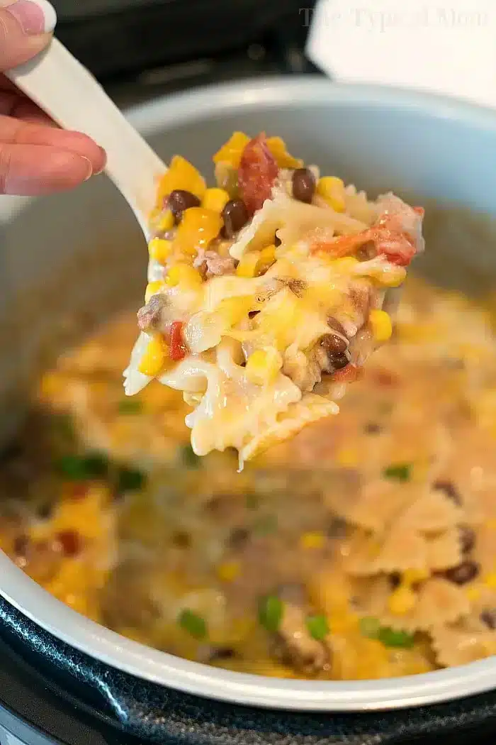 A spoonful of bowtie pasta mixed with corn, black beans, and melted cheese is held above the pressure cooker, showcasing a creamy Mexican casserole with diced tomatoes and green onions.