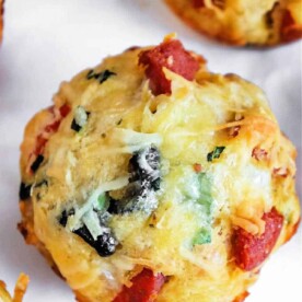 Close-up of a savory pizza muffin with visible pieces of cheese, olives, green herbs, and red ingredients, possibly pepperoni or tomatoes.