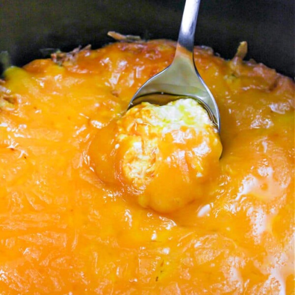 Close-up of a spoon scooping into a dish of cheesy mashed potatoes in a black slow cooker. Mimicking the ooey-gooey texture of Publix Buffalo Chicken Dip, the top layer is irresistibly covered with melted cheddar cheese.