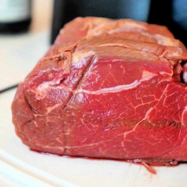 A close-up image of a raw beef roast destined for the air fryer, resting on a white surface. The marbled texture and vibrant red color of the meat are prominently displayed against a dark, blurred background.