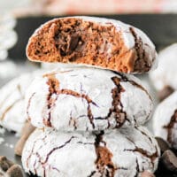 Three brownie mix crinkle cookies are stacked on a dark surface, with the top cookie broken in half to reveal the gooey chocolate inside. The cookies are dusted with powdered sugar, and chocolate chips are scattered around them.