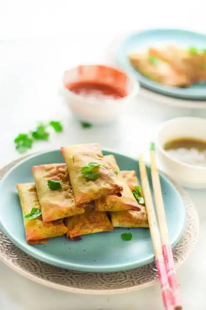 A plate with air fryer egg rolls garnished with cilantro sits on a patterned dish. Chopsticks rest beside the rolls. Two bowls, one with reddish sauce and the other with brown sauce, are in the background on a white surface.