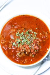 A bowl of Crockpot Venison Chili with minced meat, beans, and spices, garnished with chopped herbs, sits invitingly on a white plate accompanied by a spoon.