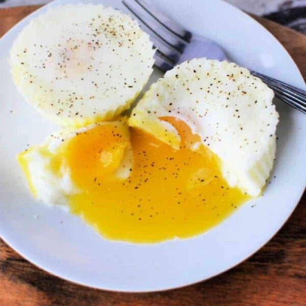 Two poached eggs on a white plate, with one cut open to reveal runny yolk spilling out, sprinkled with black pepper. Perfectly tender like slow cooker eggs, a fork rests beside them.