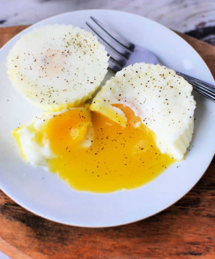 Two poached eggs on a white plate, with one cut open to reveal runny yolk spilling out, sprinkled with black pepper. Perfectly tender like slow cooker eggs, a fork rests beside them.